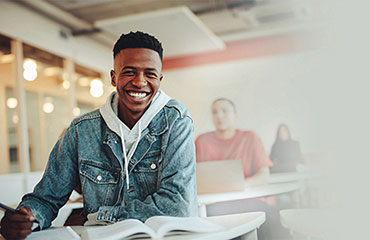 Estudiante adulto joven sentado en un aula y sonriendo con un bolígrafo en la mano y un libro abierto.