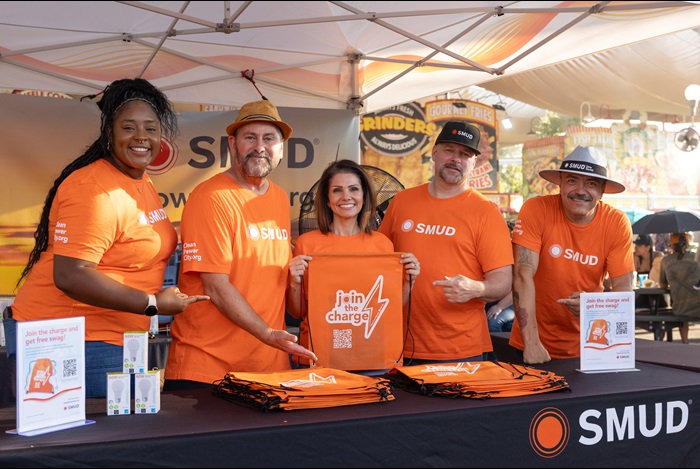 SMUD employees gathered around a SMUD booth holding up "Join the Charge" giveaway items at an outdoor event.